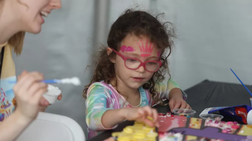 A child participates in a collaborative mosaic activity in our &quot;Kids @ Art&quot; tent, aided by a volunteer.