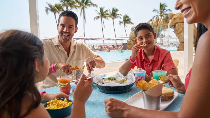 family eating lunch by pool with palm trees in the background