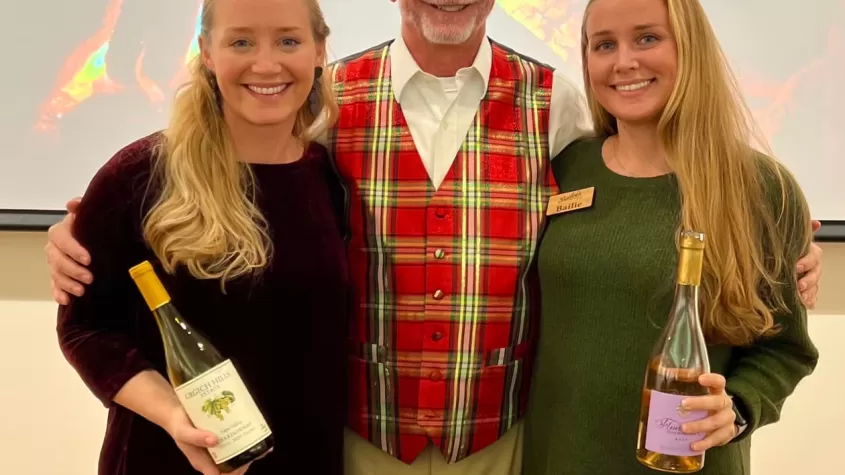 3 people stand together in festive holiday attire, holding wine bottles and glasses, with a fireplace projected onto a screen behind them 